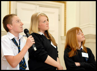 Teenager speaking into a microphone with 2 teenagers standing beside him